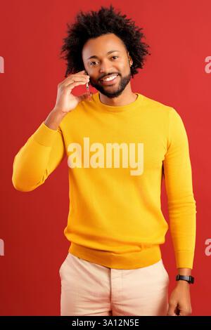 Hilarious African American curly young man talking by smartphone with empty screen and laughing isolated on red background, overjoyed guy in yellow pullover has pleasant phone call, chatting Stock Photo