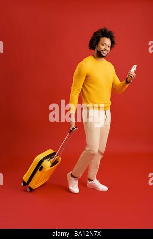 Handsome man with yellow luggage suitcase walking with smartphone to huge smartphone with empty display, copy space. Mobile app for travellers concept Stock Photo