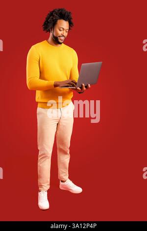 Curly male entrepreneur carrying laptop isolated on red, bearded businessman using laptop, typing, receiving emails, using new computer app, looking at screen and smiles Stock Photo