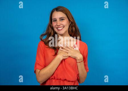 Young woman holding hands on chest, expressing gratitude, happiness, and relief, smiling on blue studio background Stock Photo