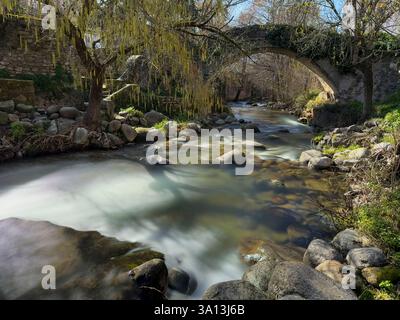 Scenic view of a river flowing under an ancient stone bridge, surrounded by lush vegetation and rocks, creating a tranquil natural landscape Stock Photo