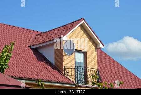 Metal roof with brick dormer, window, metal balcony, satellite dish, gutter, plastic soffits against blue sky Stock Photo