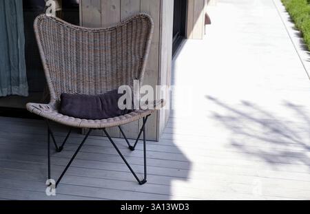 Stylish wicker chair with a soft pillow on a wooden terrace. Sunlight creates warm shadows, adding a cozy and modern ambiance to this outdoor relaxati Stock Photo