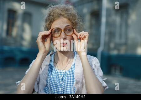 Young woman with freckles adjusting oversized vintage glasses, looking curiously at the camera, wearing a knitted sweater and checkered dress outdoors Stock Photo