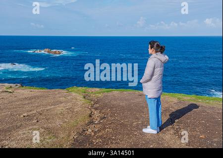 A woman wearing a gray winter coat and jeans stands on the edge of a coastal cliff, gazing at the vast blue ocean. The deep blue waves crashing Stock Photo