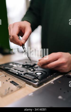 Vertical cropped shot of electronics repair professional carefully disassembling laptop motherboard, precision screwdriver in hand, surrounded by Stock Photo