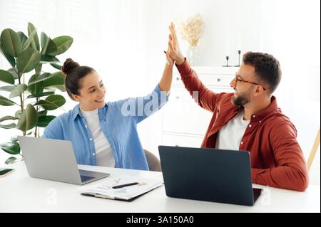 Good team job. Cheerful a man and a woman high five while sitting at their desk in a modern office, celebrating a success together, both are working on their laptops and enjoying their time at work Stock Photo