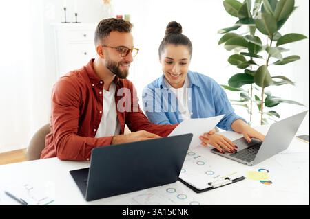 Two positive colleagues sit at their desks in an office, both working on laptops and smiling, they analyzing documents and showing them to the other, discussing work-related matters Stock Photo