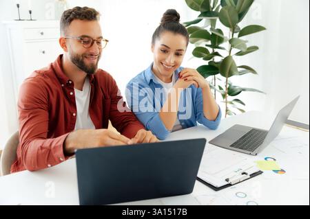 Working process of two successful colleagues working together in an office, sitting at a desk with laptops open, reviewing documents, smiling and collaborating on a new project Stock Photo