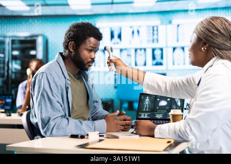 Black practitioner takes the temperature during routine checkup at the facility, using a digital thermometer to check for flu symptoms. Health physician ensures diagnosis for a young patient. Stock Photo