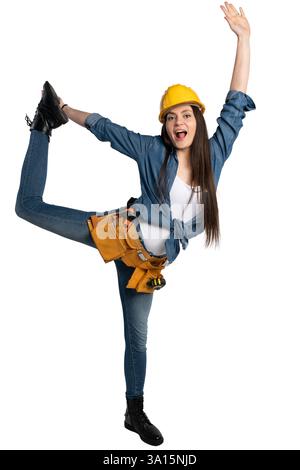 Young female construction worker wearing a yellow hard hat and tool belt, playfully lifting her leg while smiling. She is dressed in a denim shirt and Stock Photo