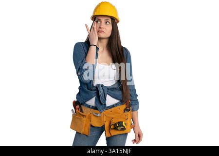 Young female construction worker wearing a yellow hard hat and tool belt, wiping her face with one hand. She is dressed in a denim shirt and jeans, ex Stock Photo