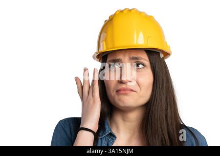 Close-up of a young female construction worker wearing a yellow hard hat, touching her face with a worried or tired expression. She is dressed in a de Stock Photo