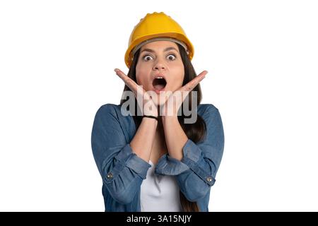 Young female construction worker wearing a yellow hard hat, holding her hands to her face with a surprised expression. She is dressed in a denim shirt Stock Photo
