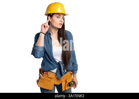 Young female construction worker wearing a yellow hard hat and tool belt, gesturing to listen. She is dressed in a denim shirt and jeans, standing aga Stock Photo