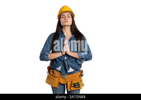 Young female construction worker wearing a yellow hard hat and tool belt, standing with hands in a meditation pose. She is dressed in a denim shirt an Stock Photo