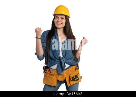 Young female construction worker wearing a yellow hard hat and tool belt, smiling confidently with raised fists. She is dressed in a denim shirt and j Stock Photo