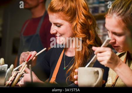 Potters working carefully on clay mugs in pottery workshop, using wooden tools Stock Photo