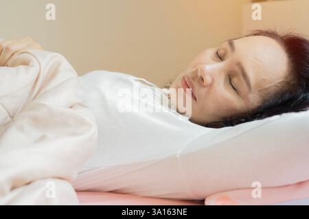 Asian mature woman sleeping in bed under white quilt with head on pillow. Close-up, side view. Stock Photo