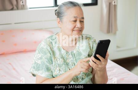 Portrait attractive mature old woman using, texting, reading message smartphone while sitting on bed in bedroom at morning. Stock Photo