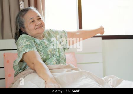 Refreshed elderly 60s female wakes up in morning stretches seated in bed in light bedroom at home. Selective focus. Stock Photo