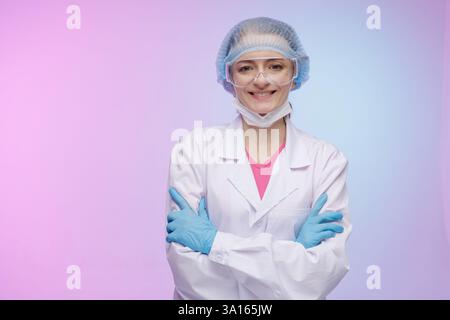 Smiling female healthcare professional in protective gear, wearing a lab coat, gloves, face mask, and safety goggles, standing confidently with arms c Stock Photo