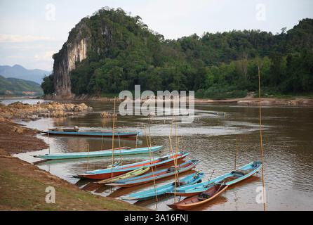 Laos, Mekong, Pak Ou caves, colourful fishing boats lined up on one bank of the river facing caves hidden in a cliff covered in vegetation Stock Photo