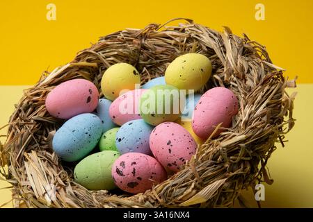 A nest made of straw is filled with assorted pastel colored easter eggs with dark speckles. Shot on a bright yellow background for contrast. Stock Photo