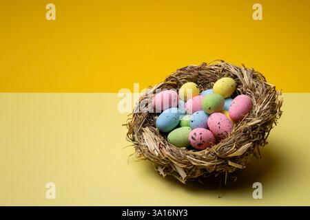 Small colorful easter eggs fill a bird's nest on a pale yellow surface with a bright yellow background, ready for spring and the easter holiday. Stock Photo