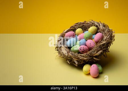 Colorful speckled easter eggs are contained in a woven nest placed on a yellow surface with a solid yellow background behind it. Stock Photo