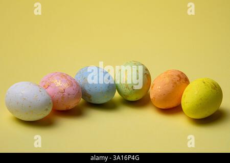 Six easter eggs in white, pink, blue, green, orange, and yellow colors with golden flecks are arranged in a line on a yellow surface. Stock Photo