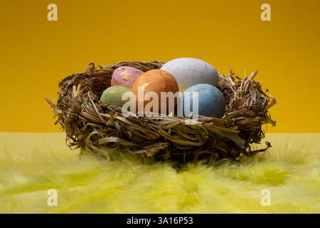 A bird's nest made of dried grass holds colorfully decorated Easter eggs. Yellow feathers are arranged beneath the nest on a yellow surface. Stock Photo