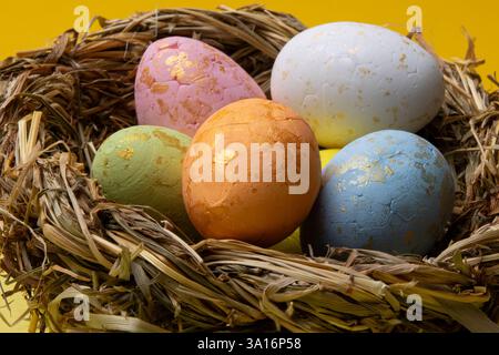 Decorated easter eggs of pink, white, green, orange, and blue colors with gold flakes are in a straw nest against a bright yellow background. Stock Photo