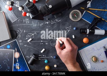 Physics student doing astronomical calculations writing with white chalk on a black board full of study material books and photographs. Top view. Stock Photo