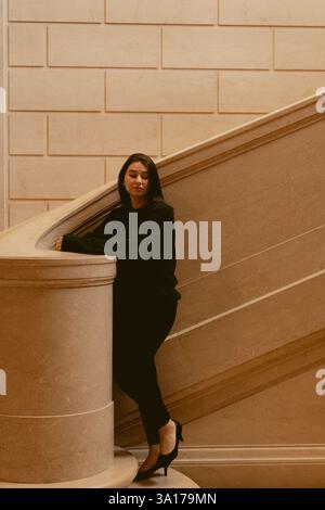 Woman in black outfit leaning on a marble staircase in soft light Stock Photo