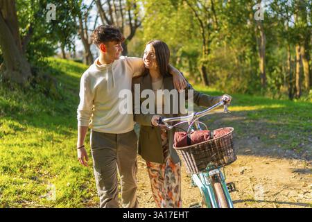 Young couple walking hand in hand through a picturesque park, riding a bicycle and basking in the warm glow of golden hour sunlight Stock Photo