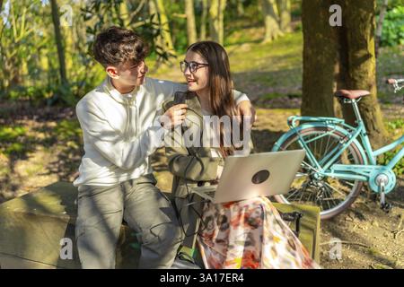 Two students are working on a laptop in a park, charging it with a power bank while sitting on a bench near their bicycle Stock Photo