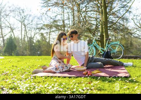 Happy couple sharing a romantic picnic in a sunny park with their bicycle nearby, enjoying fresh fruit and good company amidst blooming flowers Stock Photo