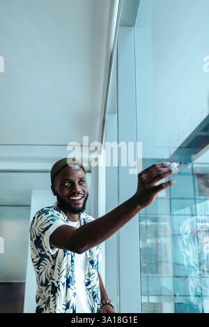 An African-American businessman, dressed casually, writes on a transparent whiteboard in a futuristic office. His confident and engaged expression ref Stock Photo