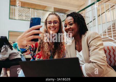Two friendly coworkers from diverse backgrounds capture a happy moment with a selfie in a modern office setting. Their joyful expressions highlight po Stock Photo