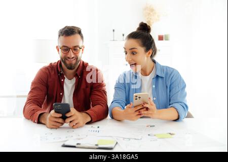 Excited man and woman sitting at a desk at office, using their smartphones, reading unexpected news, searching information, demonstrations happy emotions and amazement Stock Photo