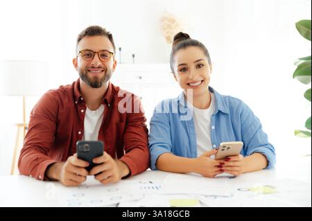 Gadgets concept. Two colleagues, man and woman sitting at a desk at office, using their smartphones, messaging online with clients or friends, reading news, searching information, smile at camera Stock Photo