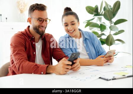 Gadgets concept. Two colleagues, man and woman sitting at a desk at office, using smartphones during working time, messaging online with clients or friends, reading news, searching information, smile Stock Photo