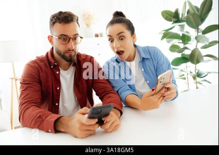 Two emotional colleagues, a man and a woman looking at their mobile phones in shocked sitting at a desk with charts and laptop, read good information, got big profit, demonstrations amazement mood Stock Photo