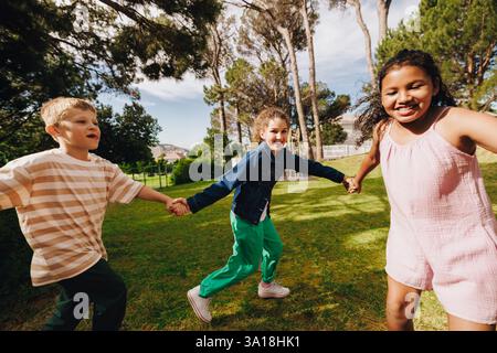 Three cheerful kids hold hands while playing together on a sunny day in a green outdoor area. The scene reflects joy, friendship, and fun activities i Stock Photo