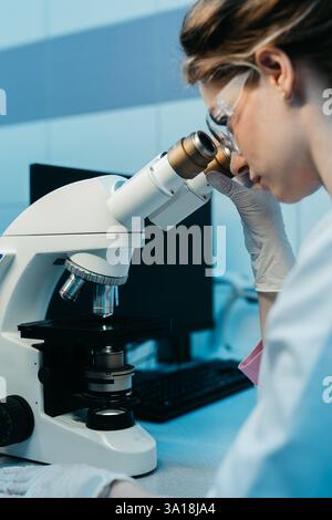A young female scientist conducts research in a medical lab, using a microscope. The concept of precise scientific experiments to discover new medical Stock Photo