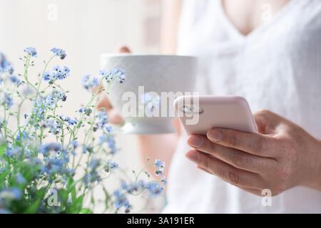 Woman's hands holding a white cup of tea or coffee and using mobile phone beside blue spring flowers Stock Photo