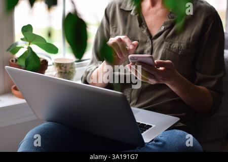 Woman sitting by a window at home with laptop computer and using mobile phone Stock Photo