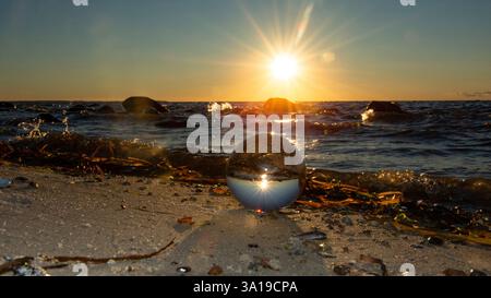 A glass ball lies in the sand in front of the sea, beach with seaweed, sea and setting sun reflected in the glass Stock Photo