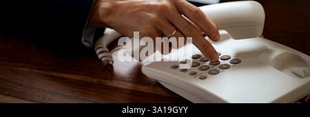 A closeup of a hand pressing buttons on a white landline telephone, with the receiver lifted. The person is wearing a ring and a dark suit. Stock Photo